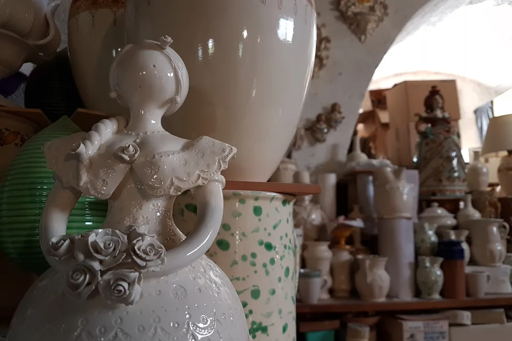 In einer Keramikwerkstatt in Grottaglie: vorn eine weiß glasierte Figur mit Keramikrosen, dahinter Regale mit Vasen und Krügen unter einem weiß gekalkten Gewölbe