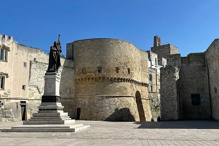 Die runde Bastion der aragonesischen Burg von Otranto über einem leeren gepflasterten Platz und einer Bronzestatue